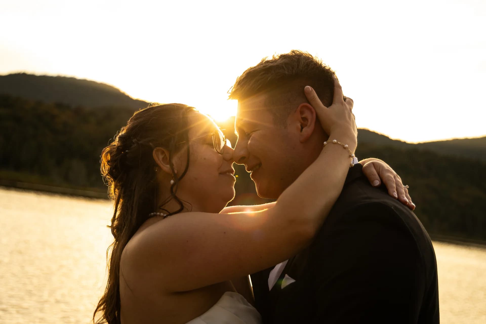 Newlyweds lean toward each other by a lake in the setting sun.